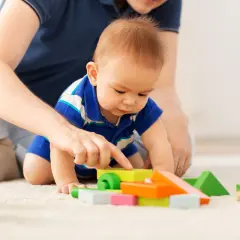 Dziecko w niebieskiej koszulce bawi się kolorowymi klockami na dywanie, a obok widać ręce dorosłego.