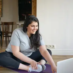 Uśmiechnięta kobieta w stroju sportowym siedzi na macie do ćwiczeń przed laptopem.