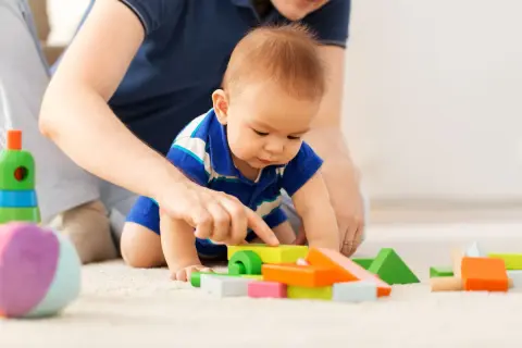 Dziecko w niebieskiej koszulce bawi się kolorowymi klockami na dywanie, a obok widać ręce dorosłego.