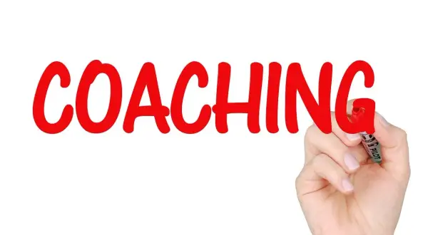 Ręka pisze czerwonym markerem słowo "COACHING" na białym tle.