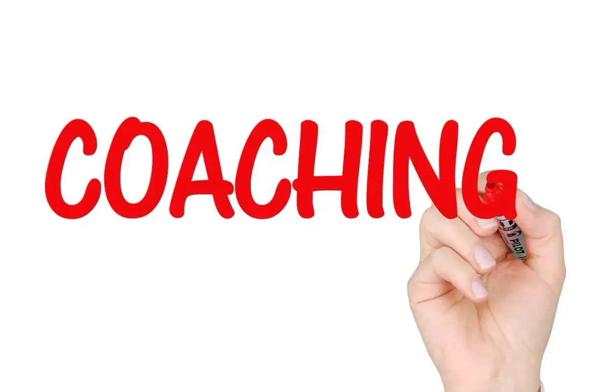Ręka pisze czerwonym markerem słowo "COACHING" na białym tle.